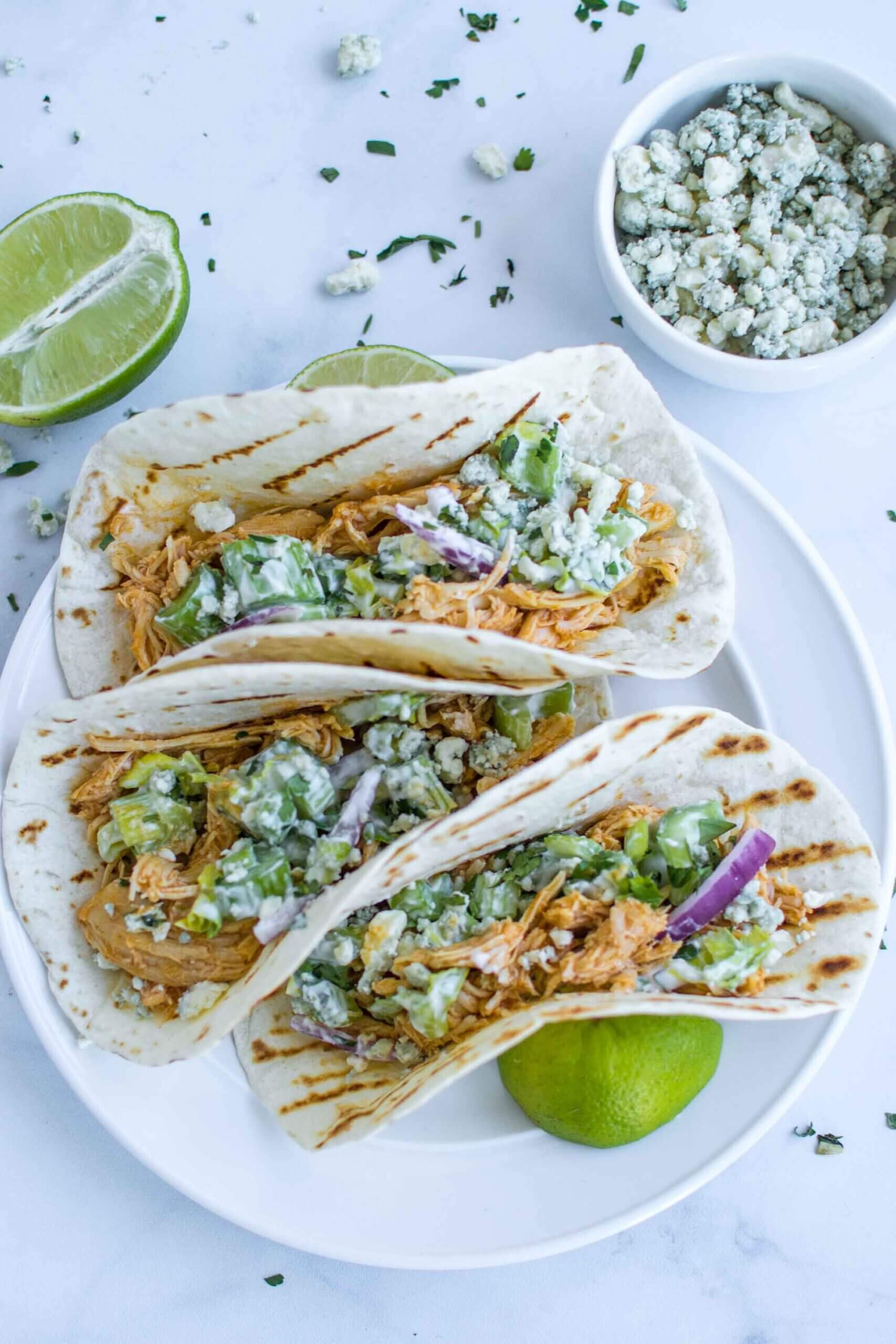 Three buffalo chicken tacos on a plate with blue cheese and lime