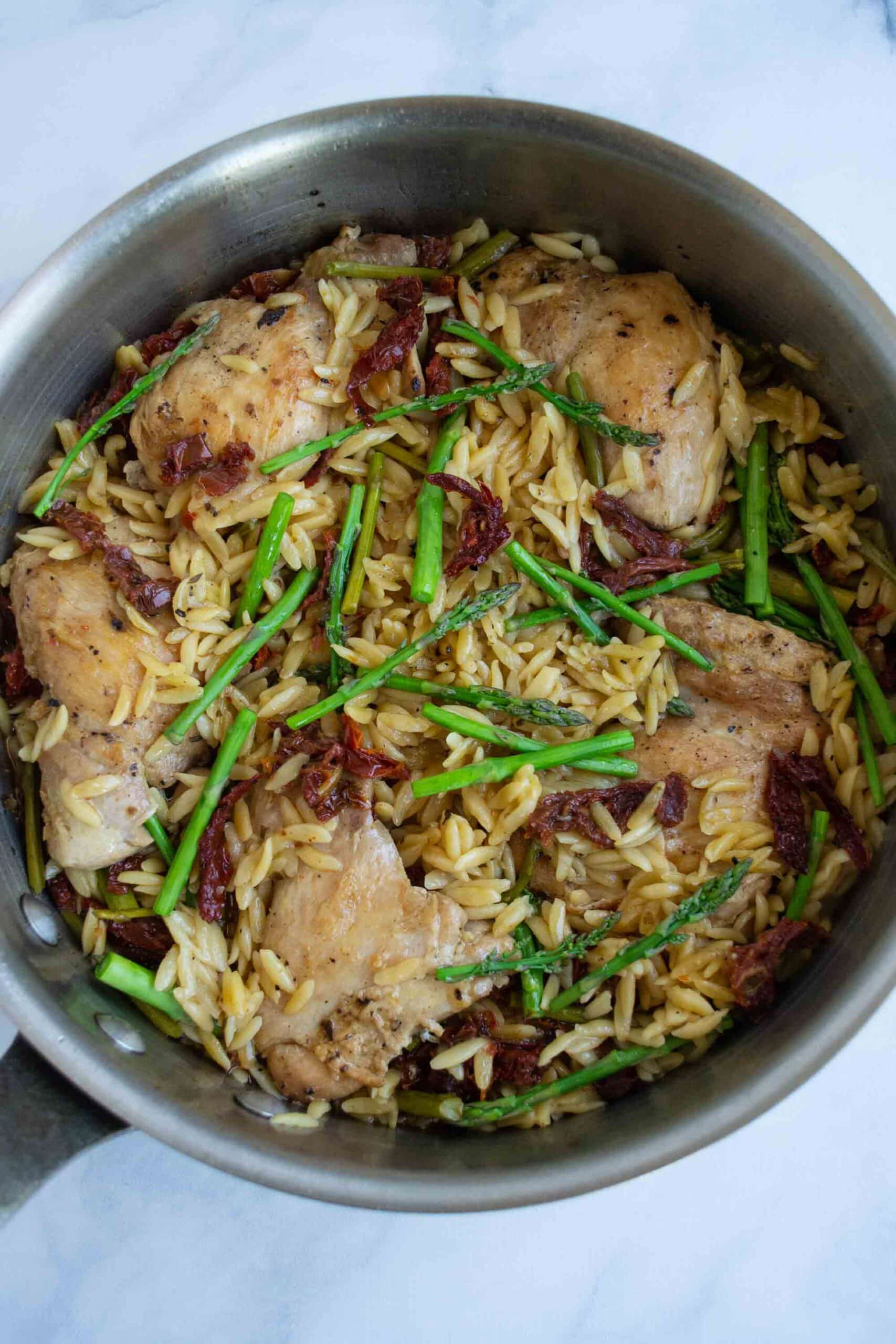 Chicken thighs with orzo in a pan with sun dried tomatoes and asparagus.