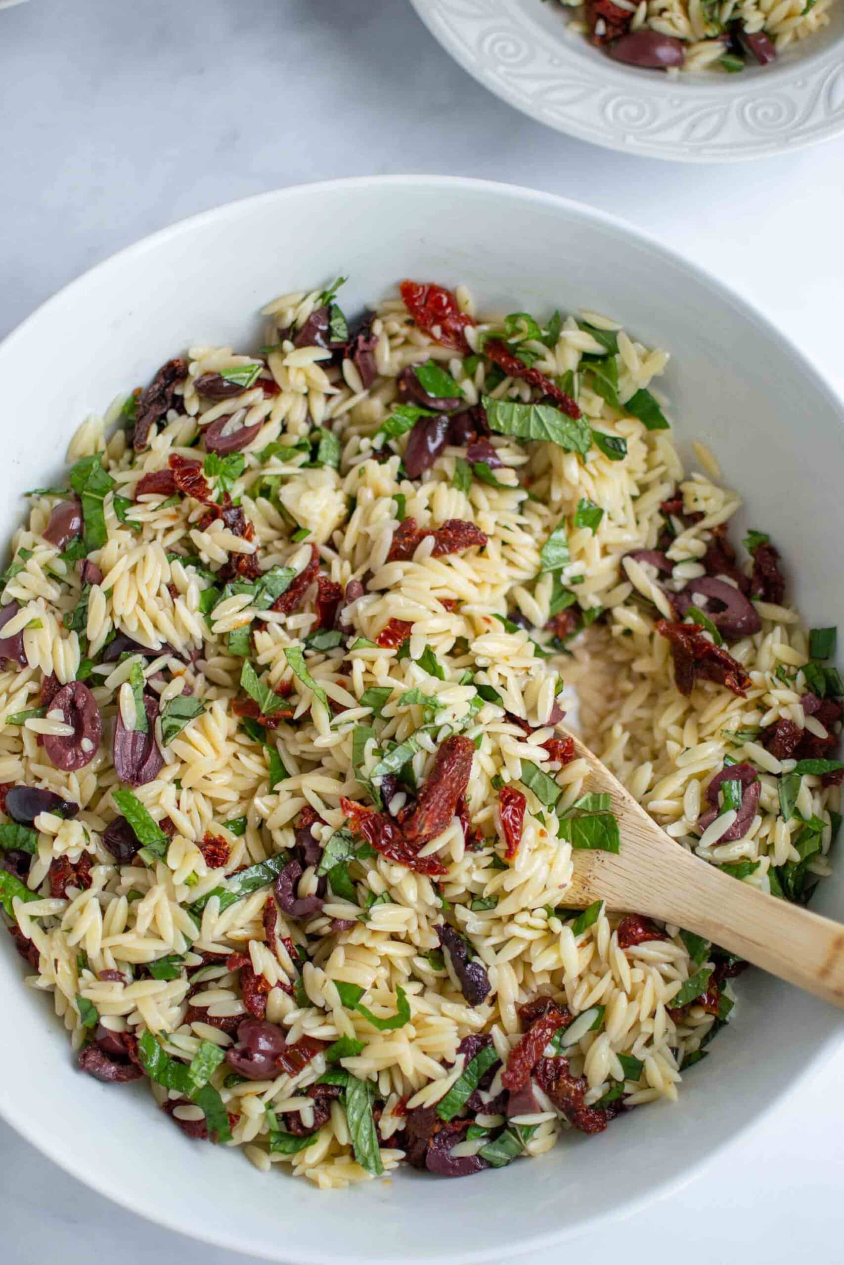 Orzo salad with olives sun dried tomatoes and fresh herbs in a large white bowl and wooden spoon.
