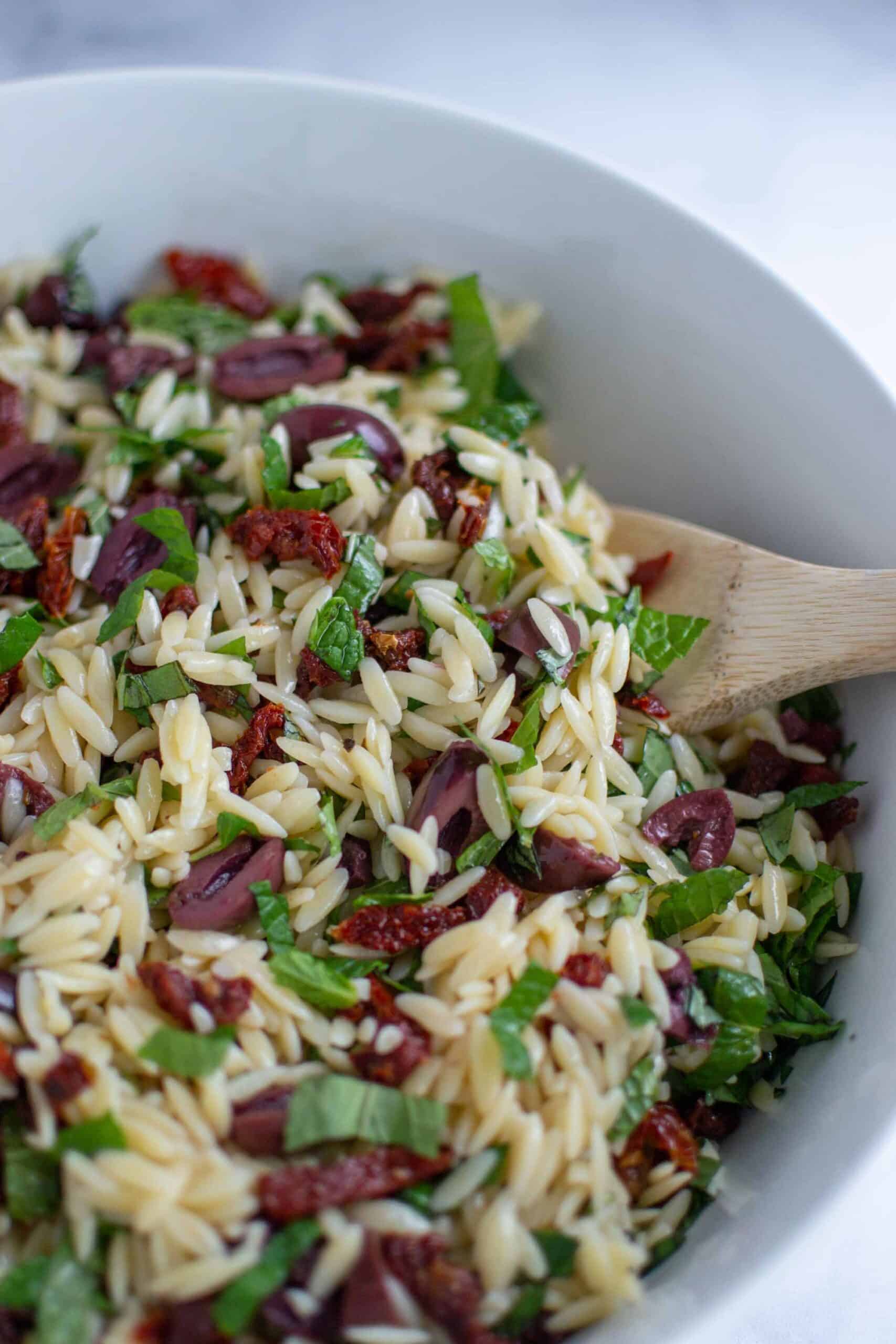 Orzo salad in a large white bowl with a wooden spoon