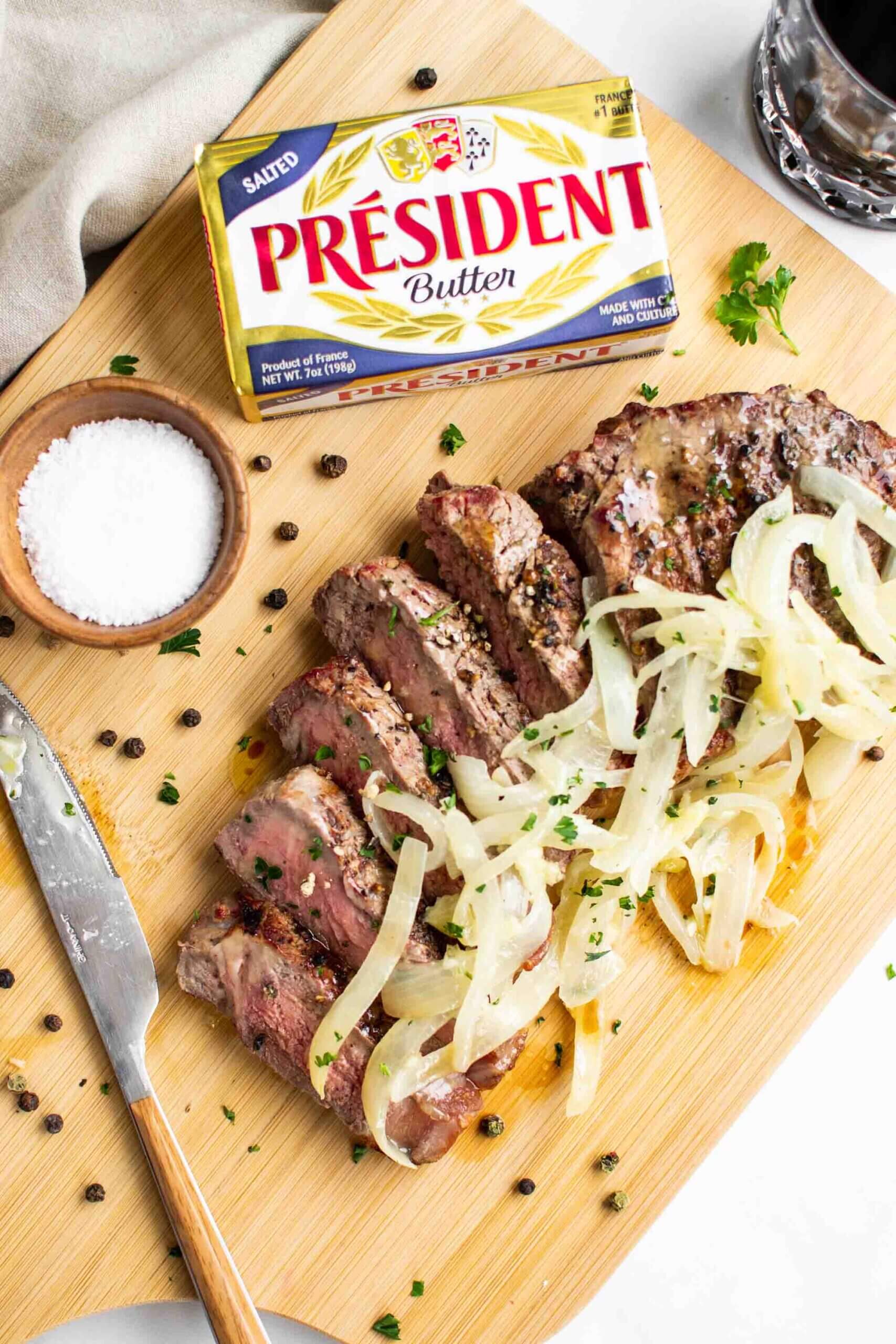 Steak with Garlic Butter Onions on a cutting board with salt and pepper and a knife and fork. Président butter block in top left.