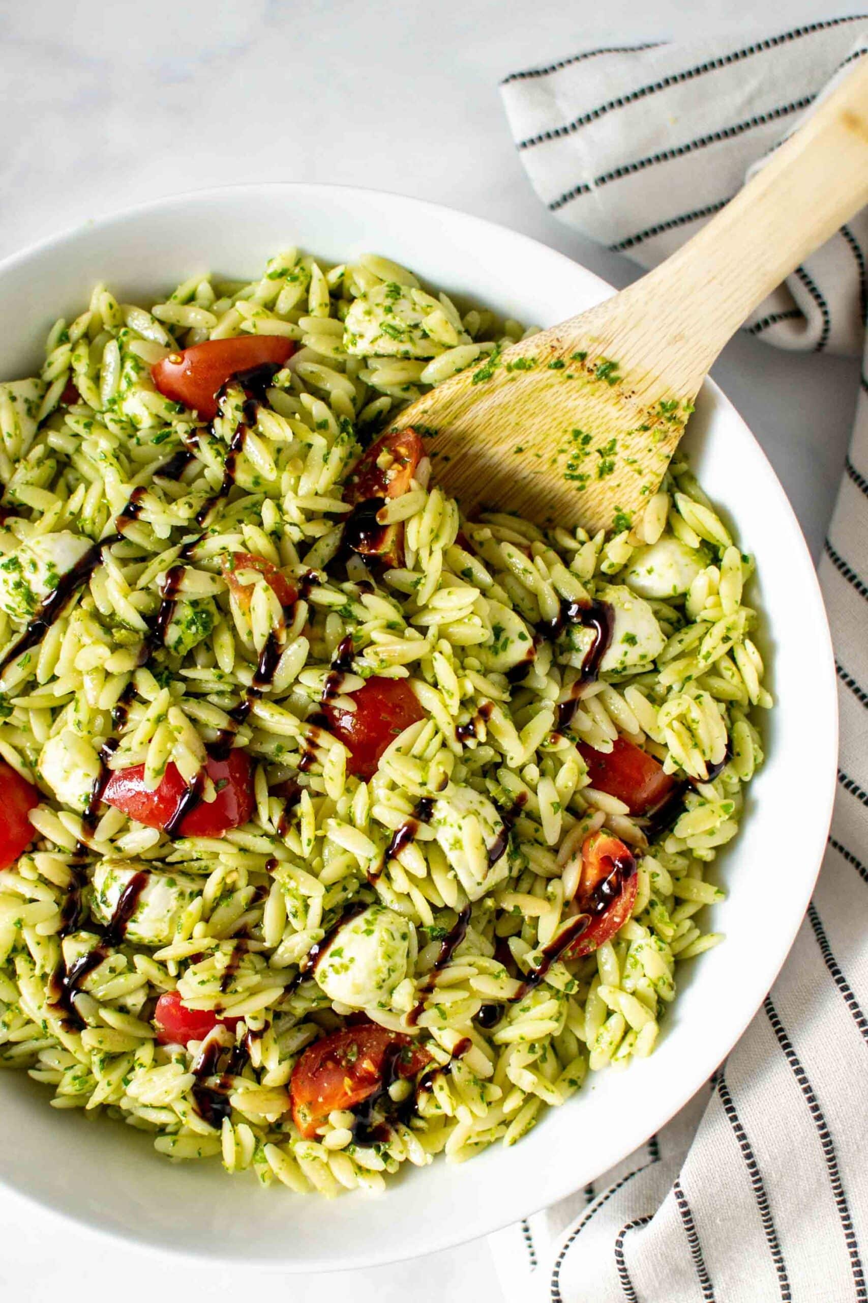 Caprese pesto orzo salad in a white bowl with a wooden spoon drizzled with balsamic glaze.