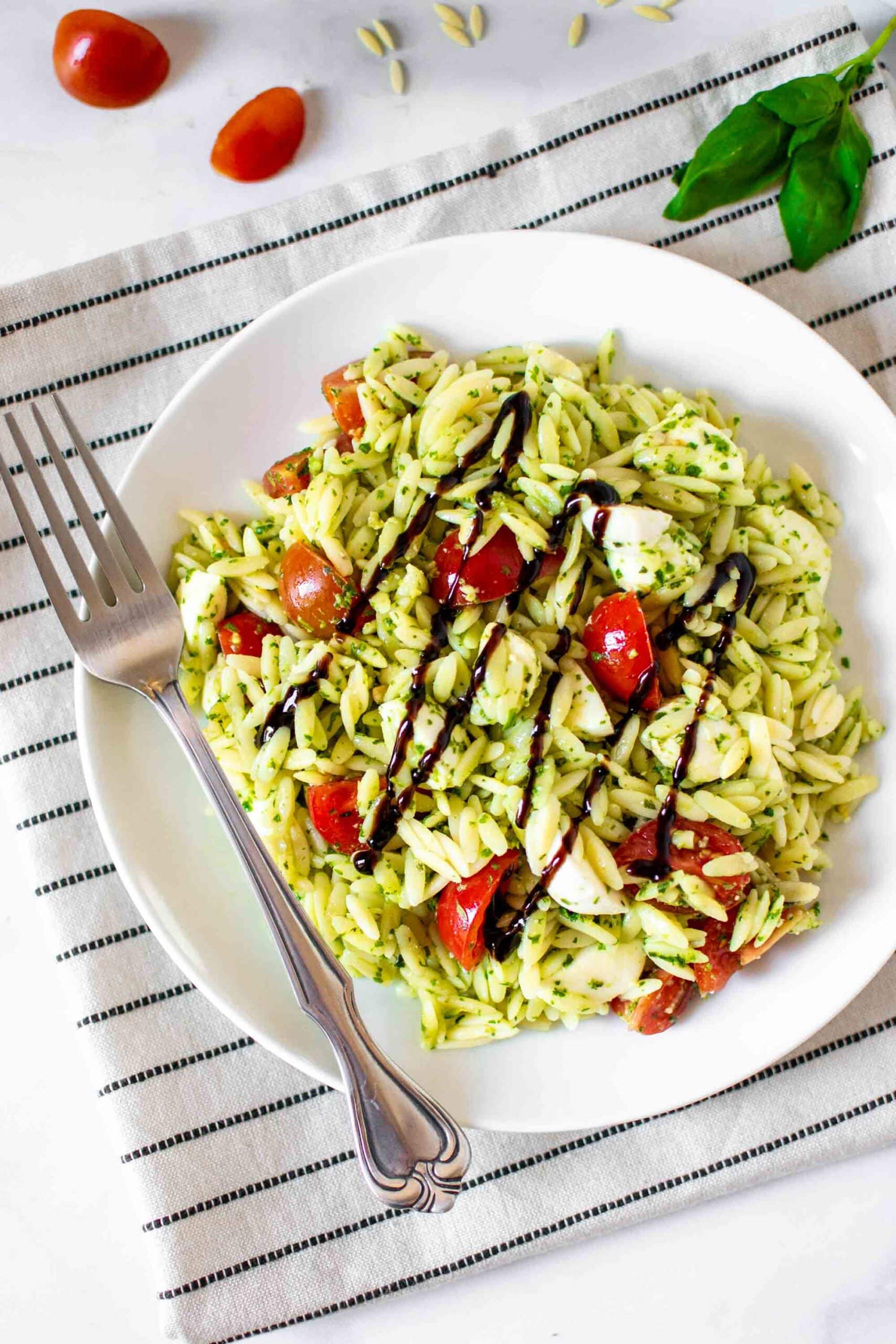Caprese Pesto Orzo Salad on a plate with fork topped with cherry tomatoes and fresh mozzarella and drizzled with balsamic glaze.