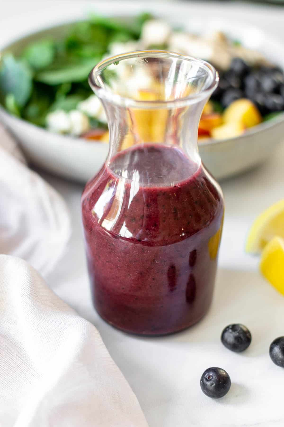 Blueberry salad dressing in a glass carafe in front of a salad.