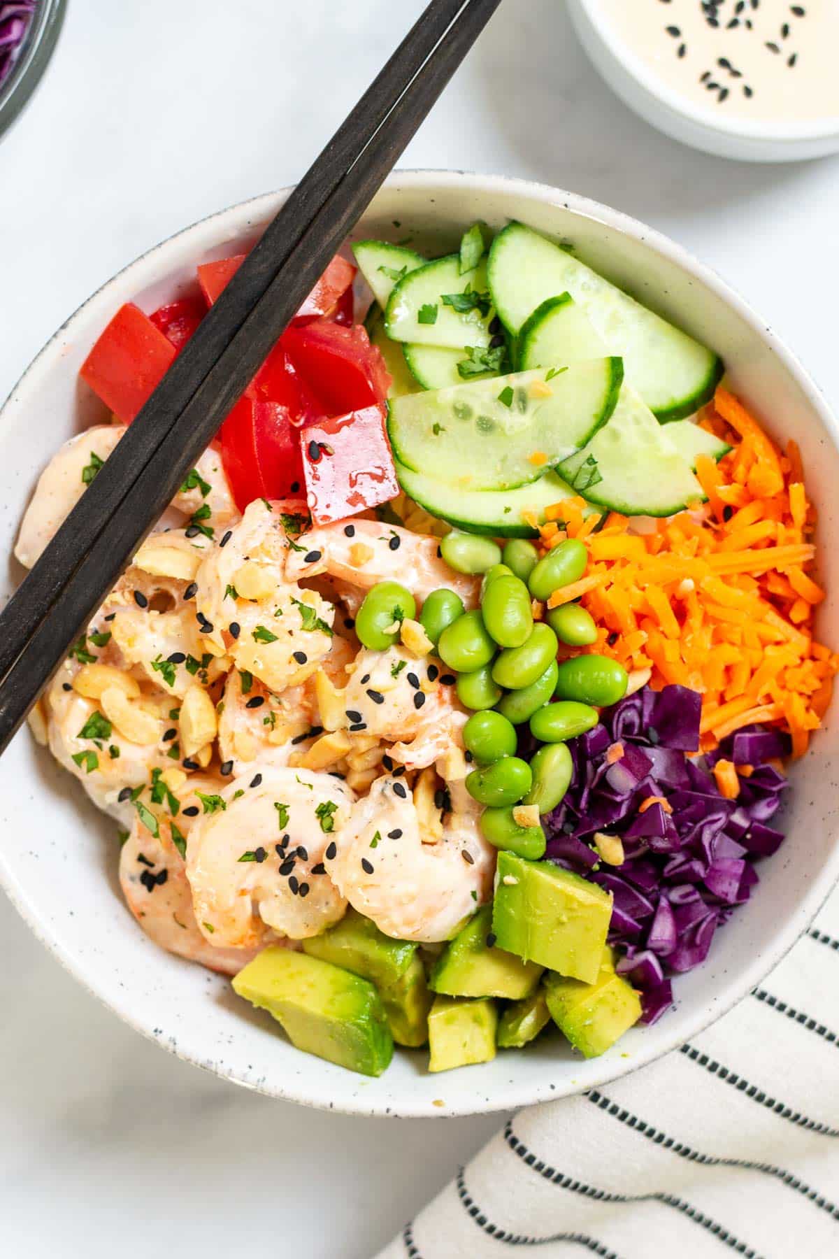 Easy shrimp poke bowl in a bowl with sriracha mayo, tomatoes, cucumber, and other veggies. Chopsticks are placed on the bowl.