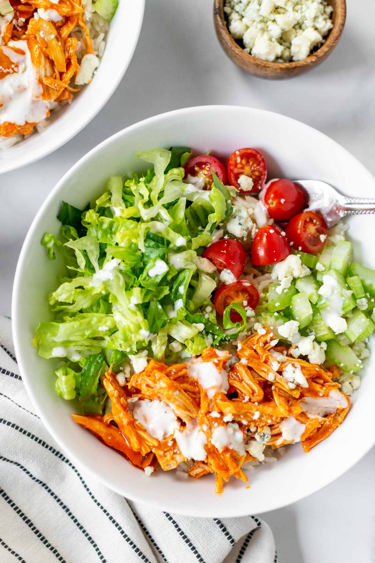 Two buffalo chicken rice bowls with a fork and a small container of blue cheese crumbles. 