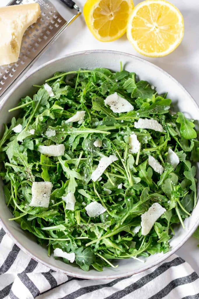 Arugula salad in a white bowl with Parmesan shavings on top.