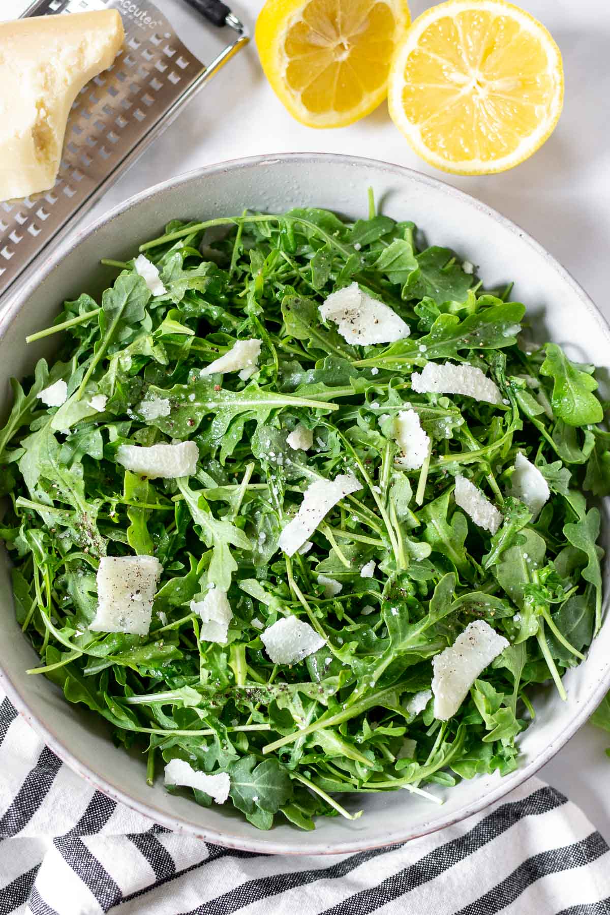 Simple arugula salad in a bowl with parmesan shavings and lemons and cheese on the side.