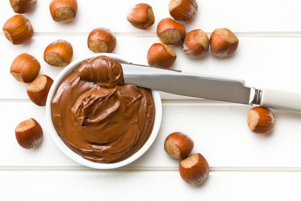 Nutella in a small white bowl with a knife and hazelnuts.