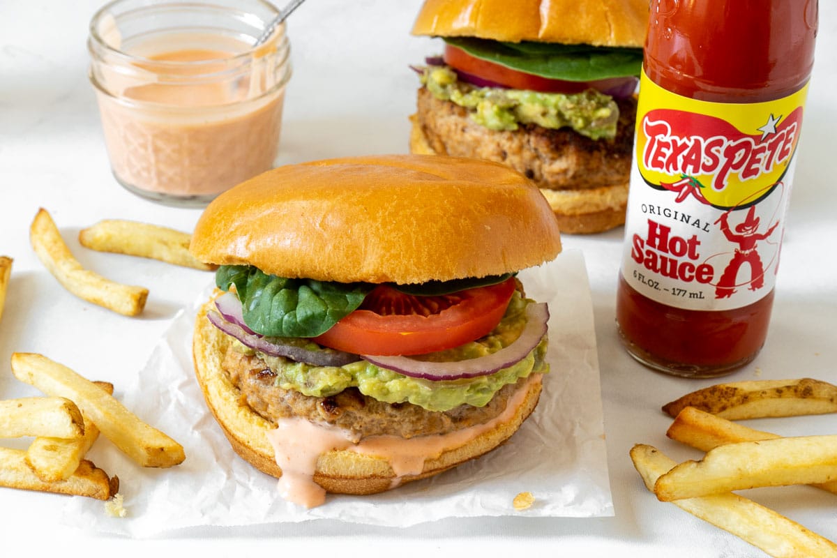 Spicy turkey burger with avocado, onion and tomato on a cutting board with Texas Pete® Hot Sauce and french fries. 
