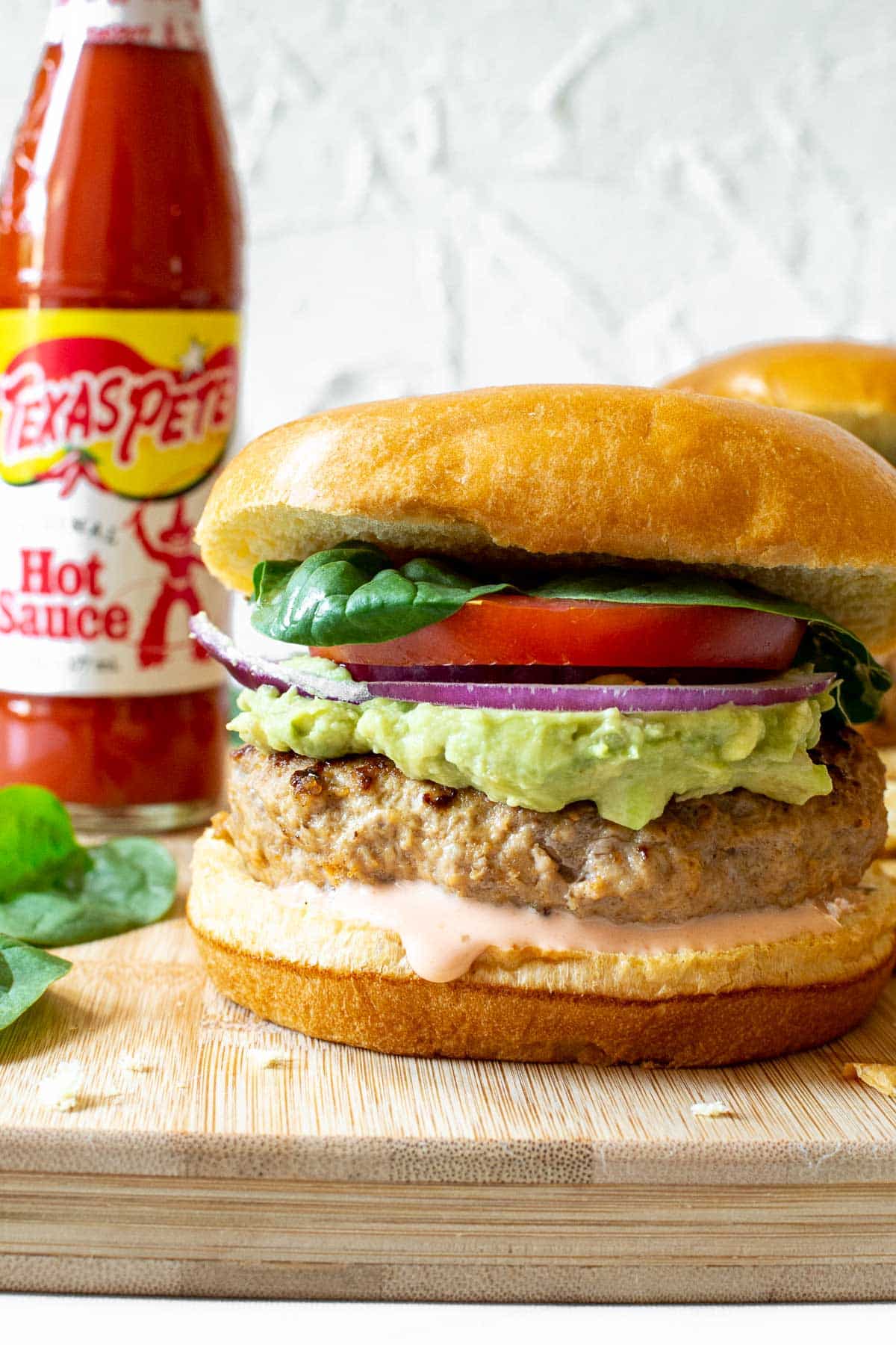 Spicy turkey burger with avocado, onion and tomato on a cutting board with Texas Pete® Hot Sauce in the background.