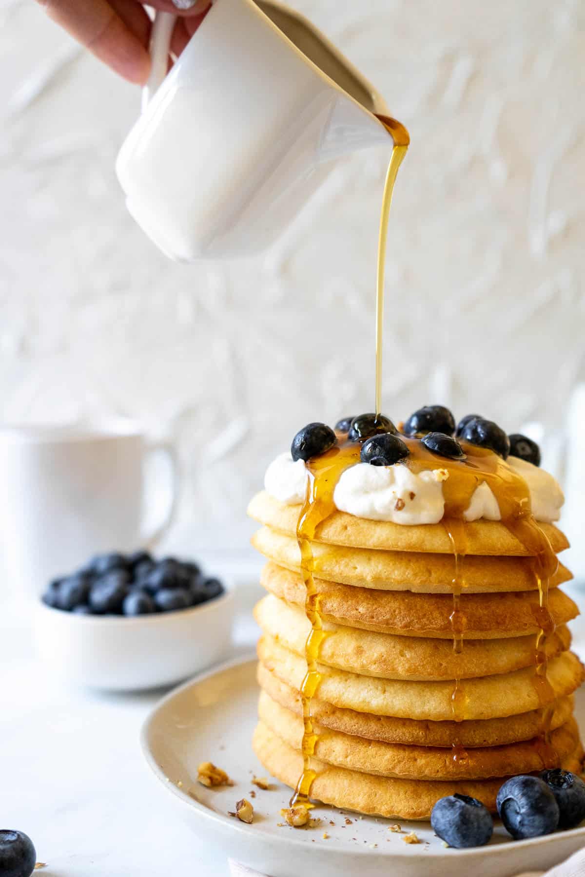 Syrup being poured onto air fryer pancakes that are topped with whipped cream and blueberries.