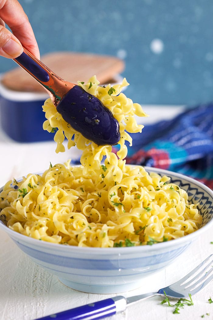 A bowl of egg noodles sprinkled with parsley being scooped up with tongs.