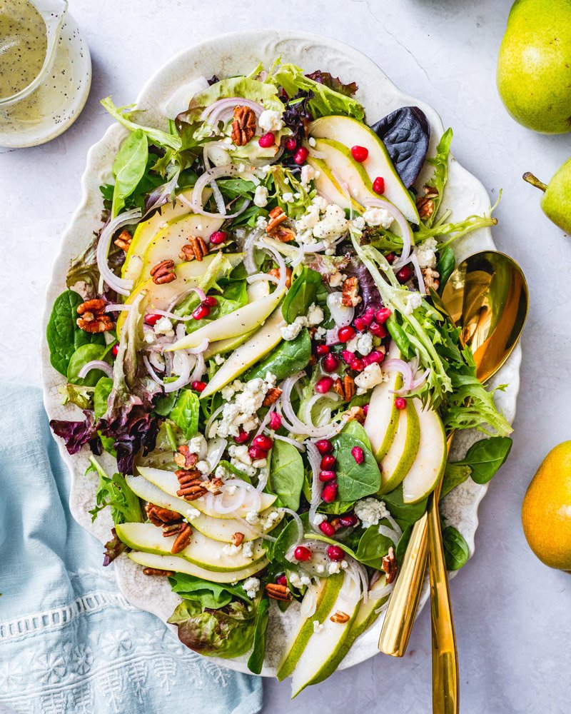 An oval dish with mixed greens, pomegranate seeds and pears. 