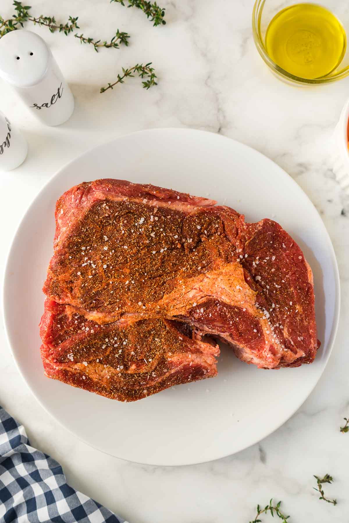 Chuck roast on a white plate being seasoned with spices.