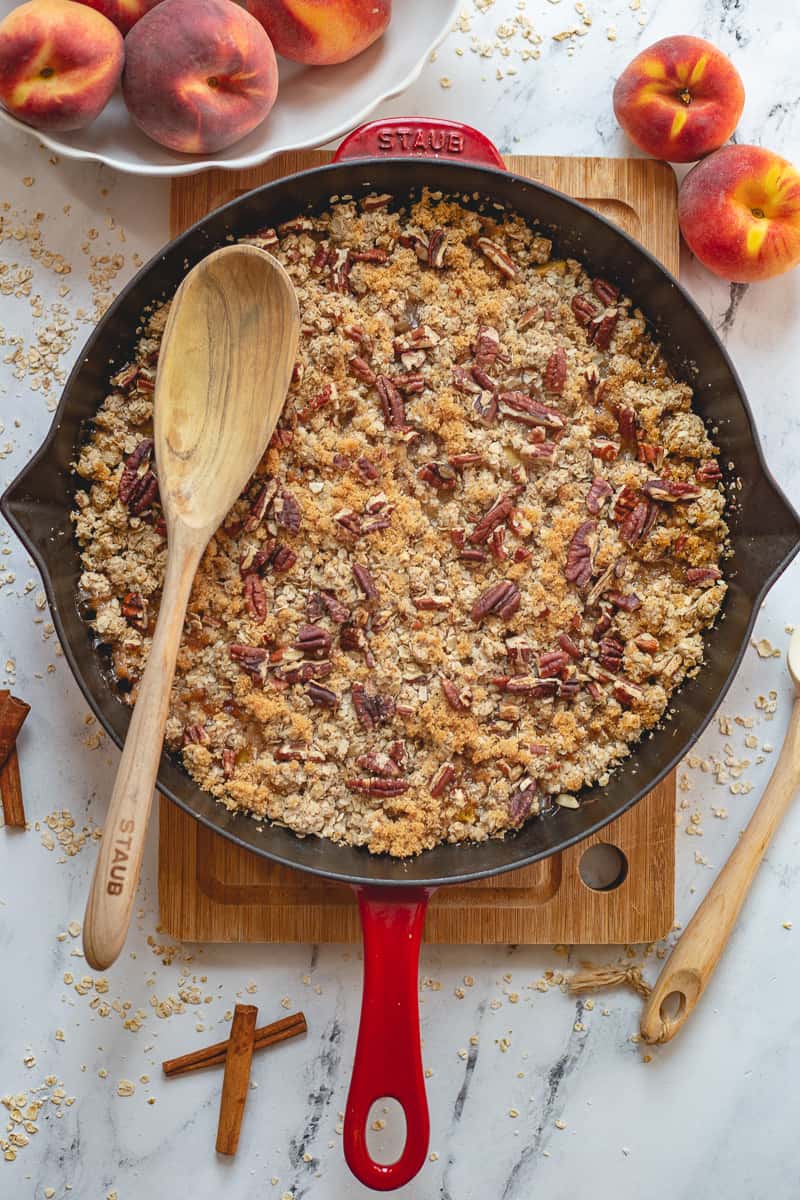 A skillet with peach crisp topped with lots of oats and pecans with a wooden spoon.