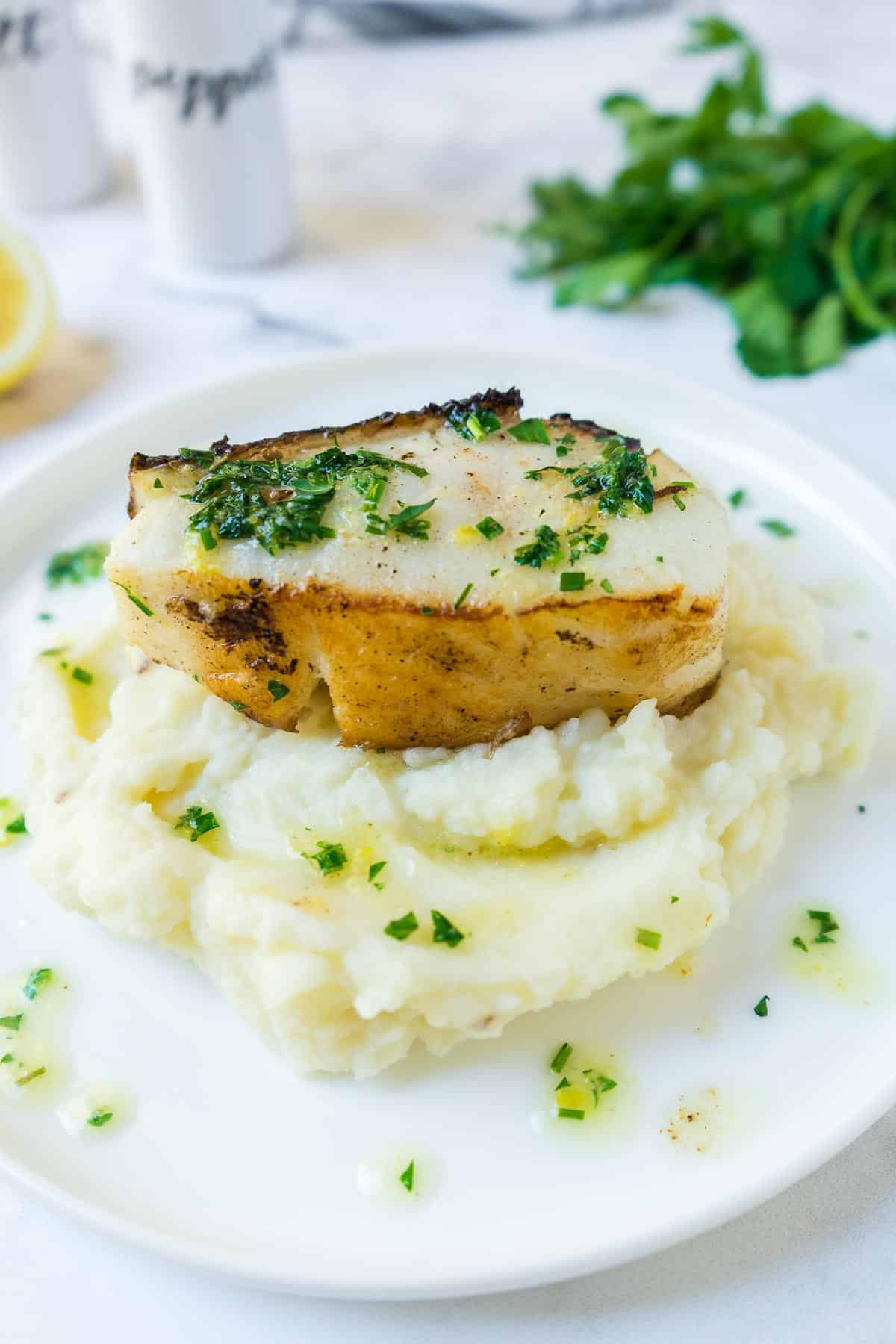Pan seared Chilean Sea Bass on a plate with mashed potatoes drizzled with the herb butter sauce.