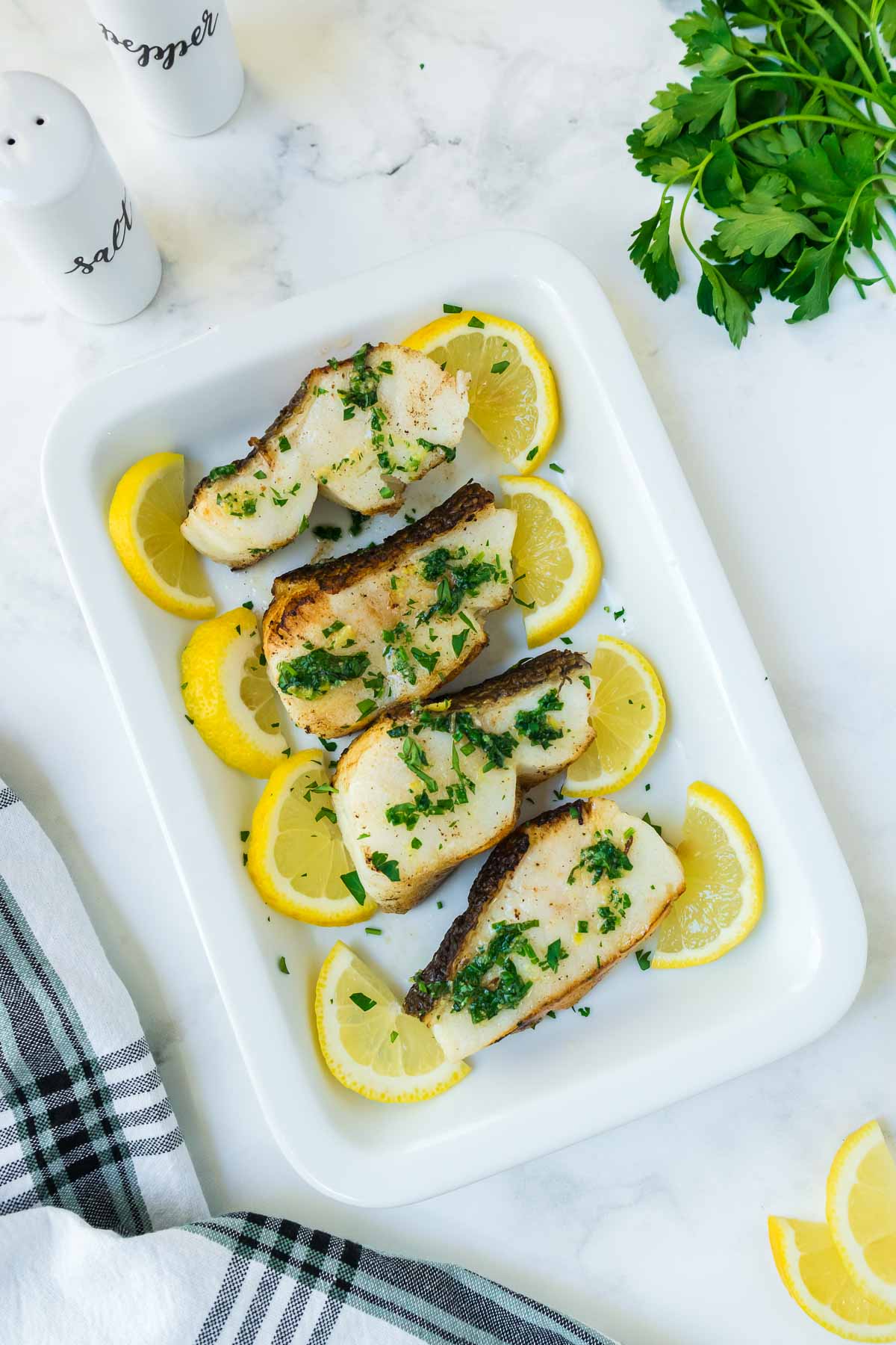 A white serving dish with cooked Chilean sea bass with herb butter sauce and lemons.
