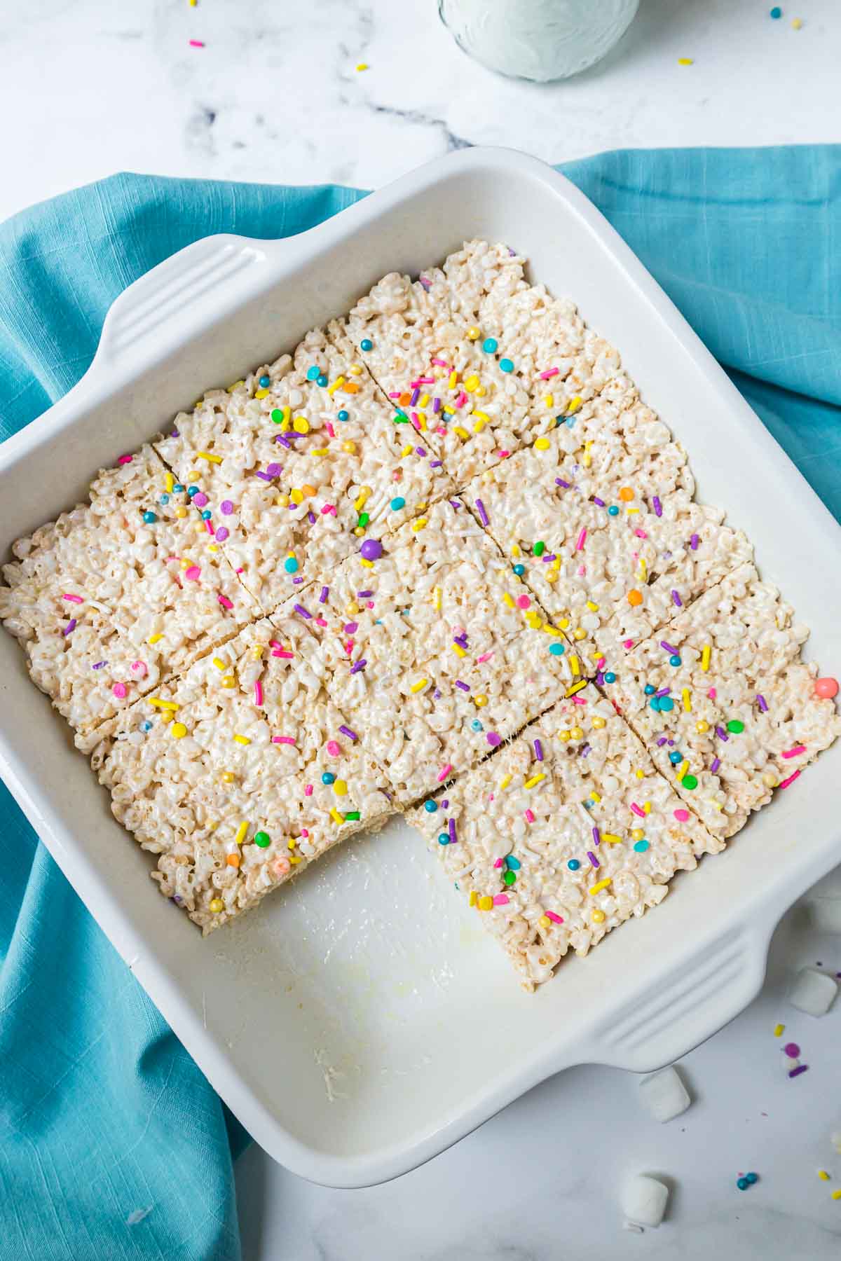 A white baking dish with rice krispies treats cut into squares with one piece missing.