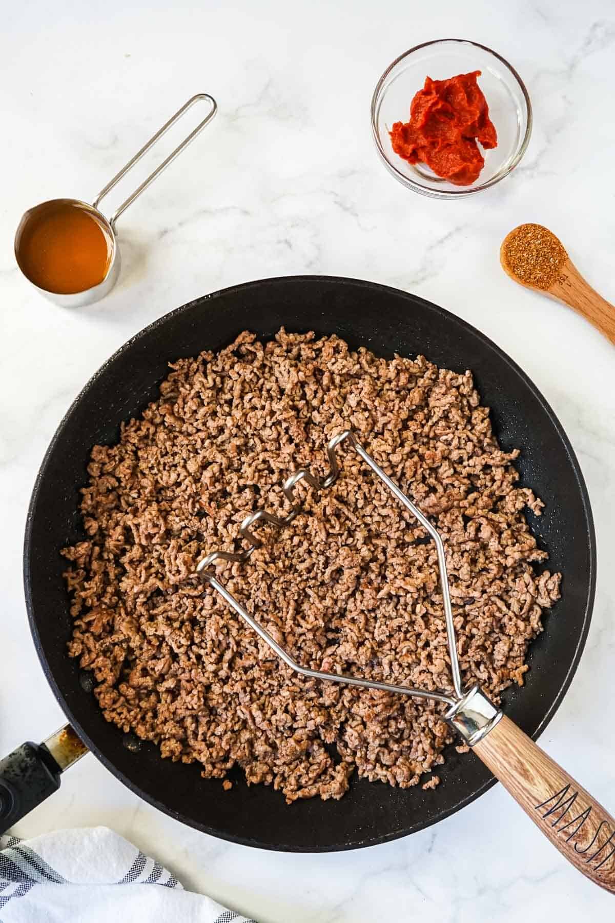 Ground beef in a skillet being cooked with a potato masher to get the meat broken up.