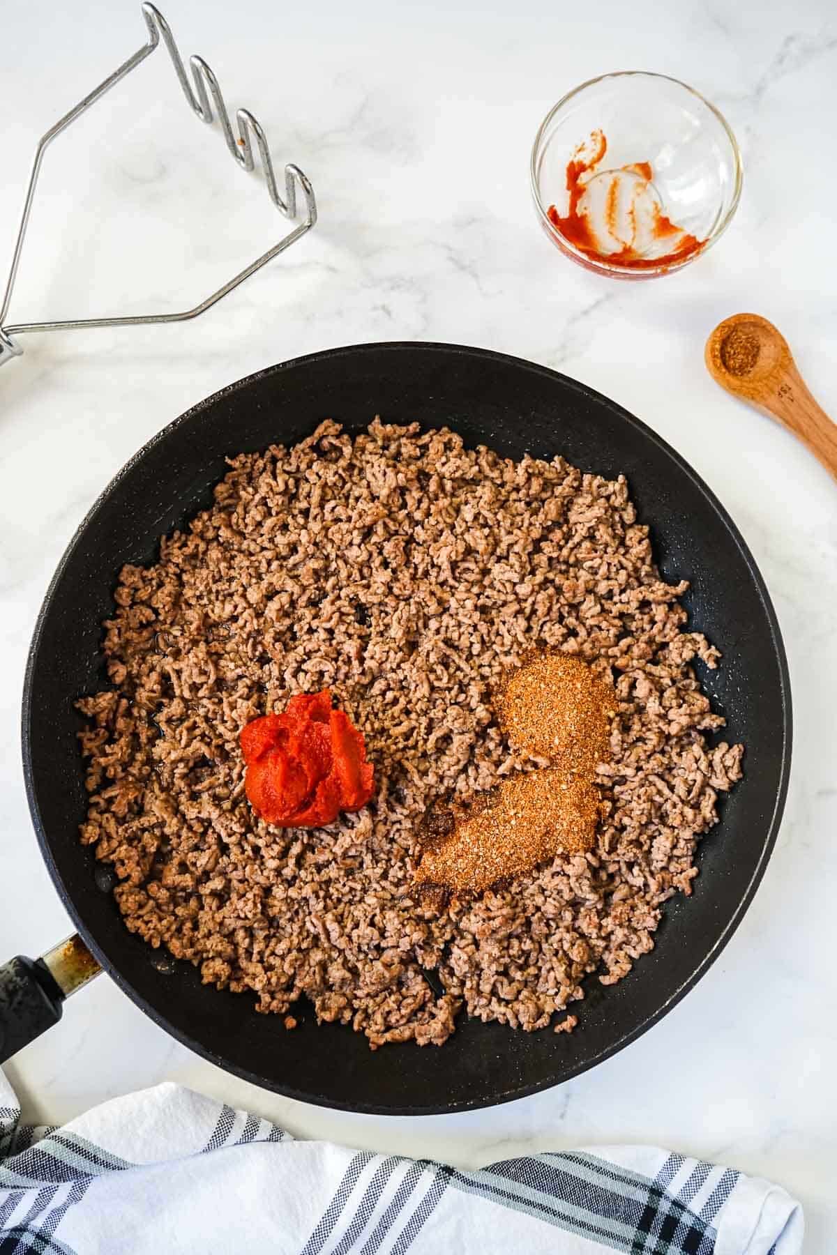 Cooked meat in a skillet with homemade taco seasoning being added along with tomato paste.