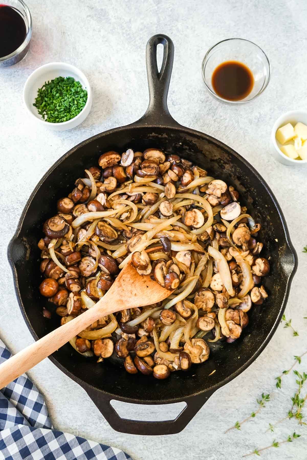 Mushrooms and onions cooking in a large cast iron skillet with a wooden spoon.