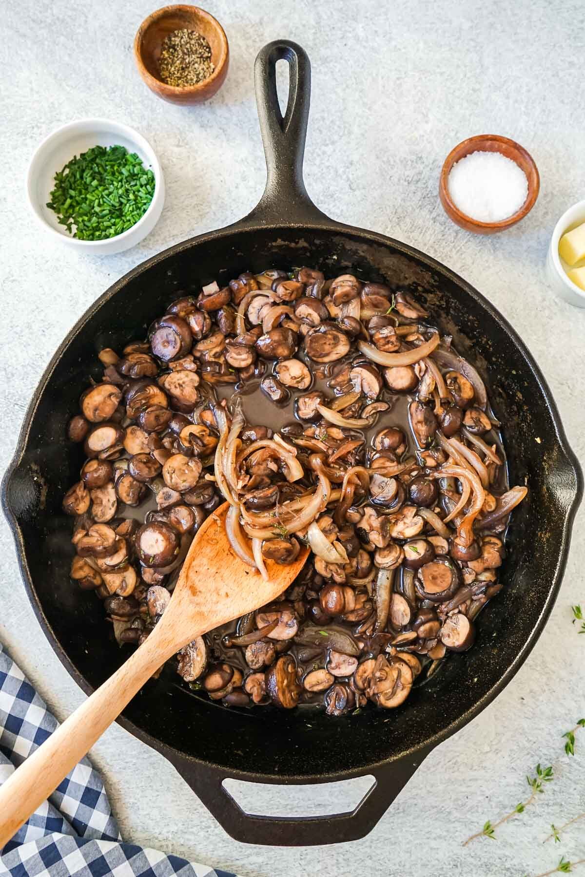 Sautéed mushrooms and onions in a cast iron skillet with a wooden spoon.