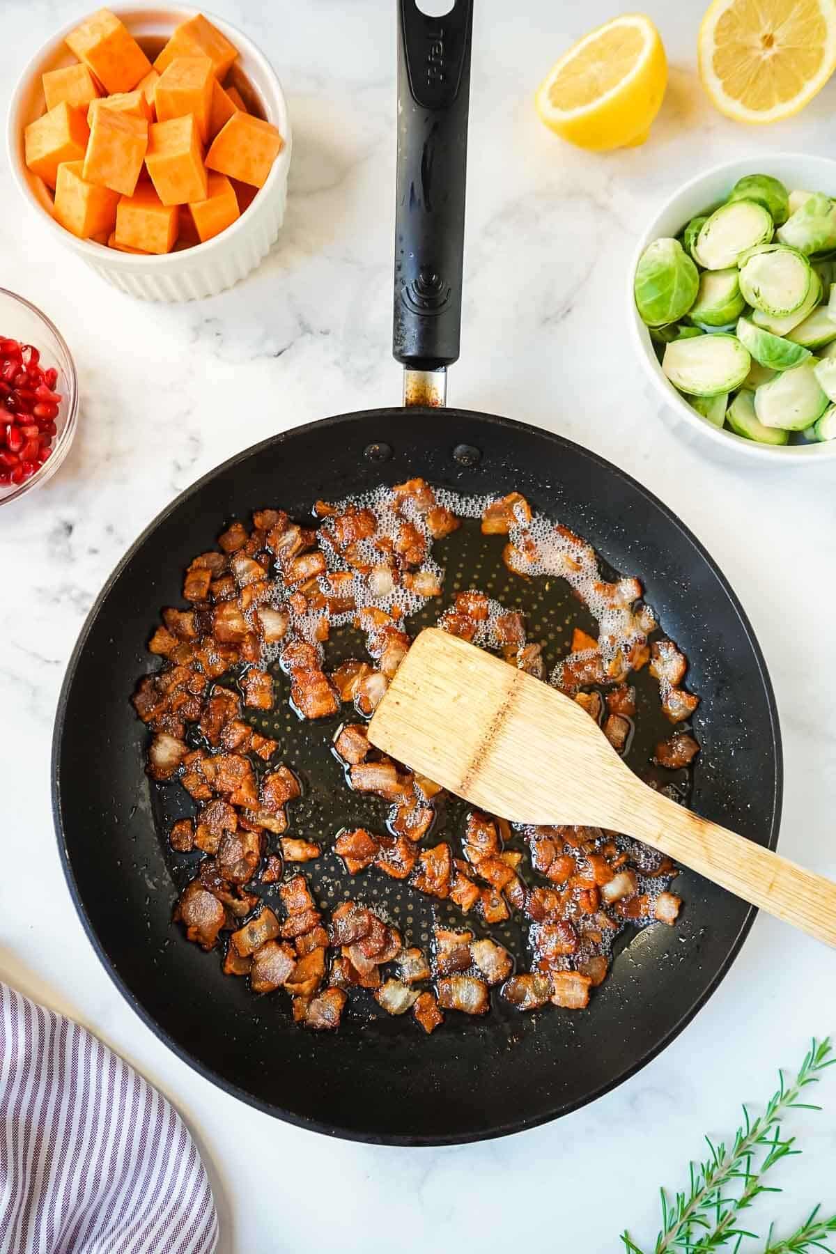 Small bacon pieces frying in a black skillet.