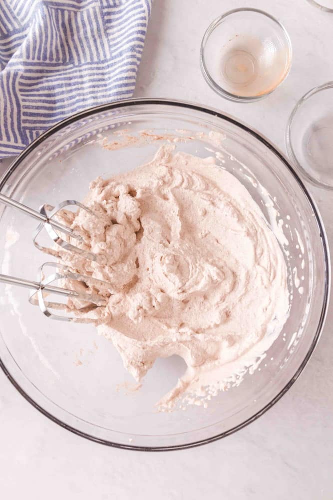 Cinnamon whipped cream in a glass bowl being beat with an electric mixer.