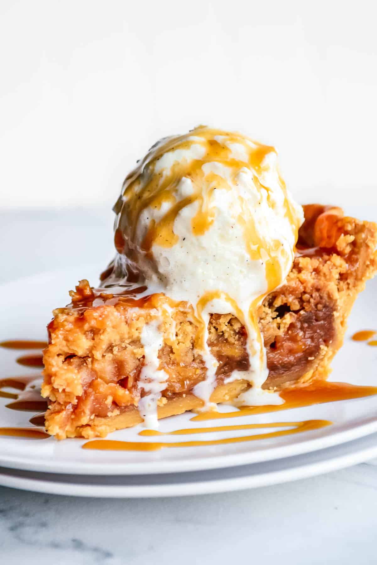 Caramel Apple Pie with Crumb Topping on a white plate with a scoop of vanilla ie cream and drizzled with caramel sauce.
