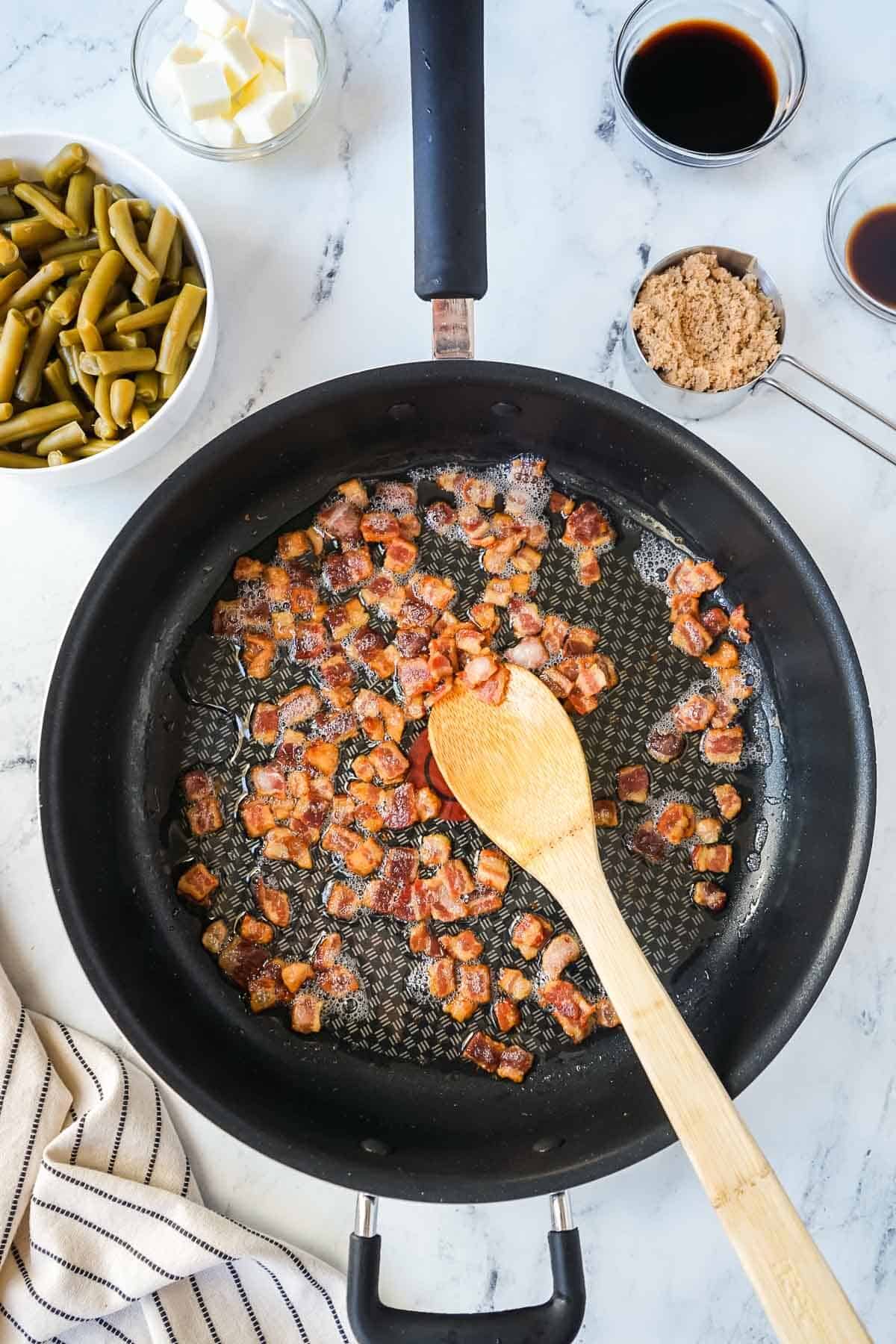 Pieces of bacon cooking in a large skillet with a wooden spoon.