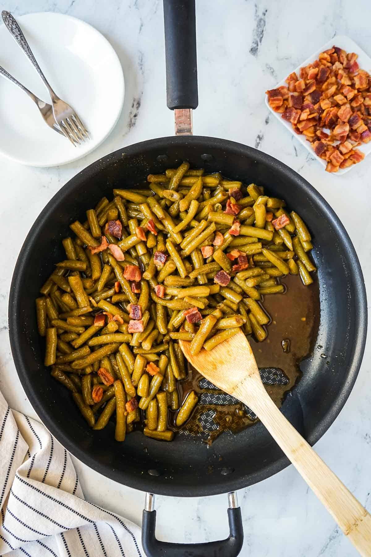 Green beans tossed with glaze and bacon in a large skillet with a wooden spoon.