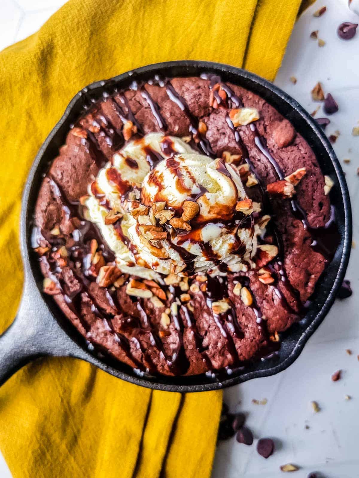 Cast iron skillet brownie with nuts and vanilla ice cream and nuts on a yellow dish cloth.