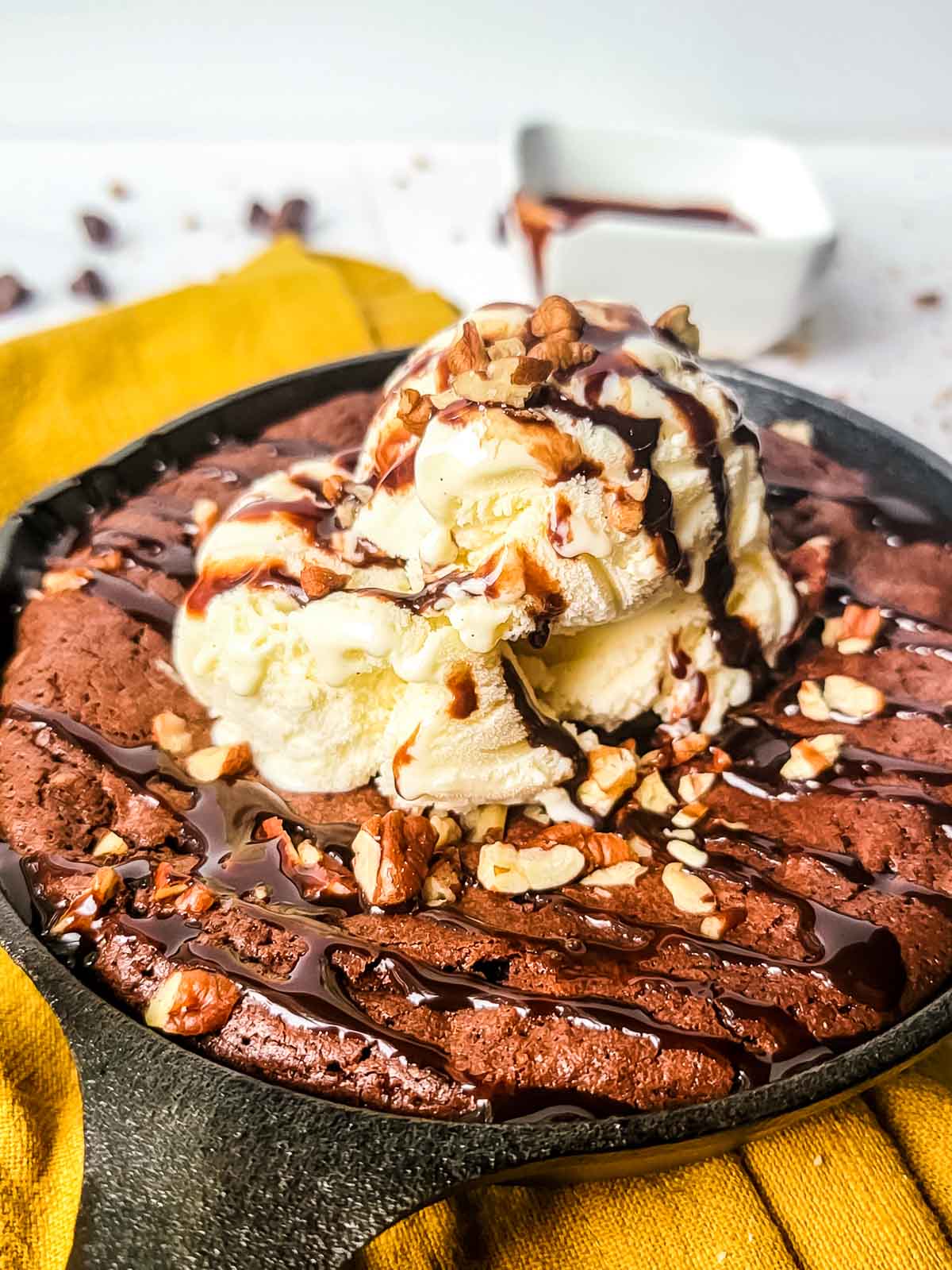 Cast iron skillet brownie with nuts and vanilla ice cream and nuts on a yellow dish cloth.