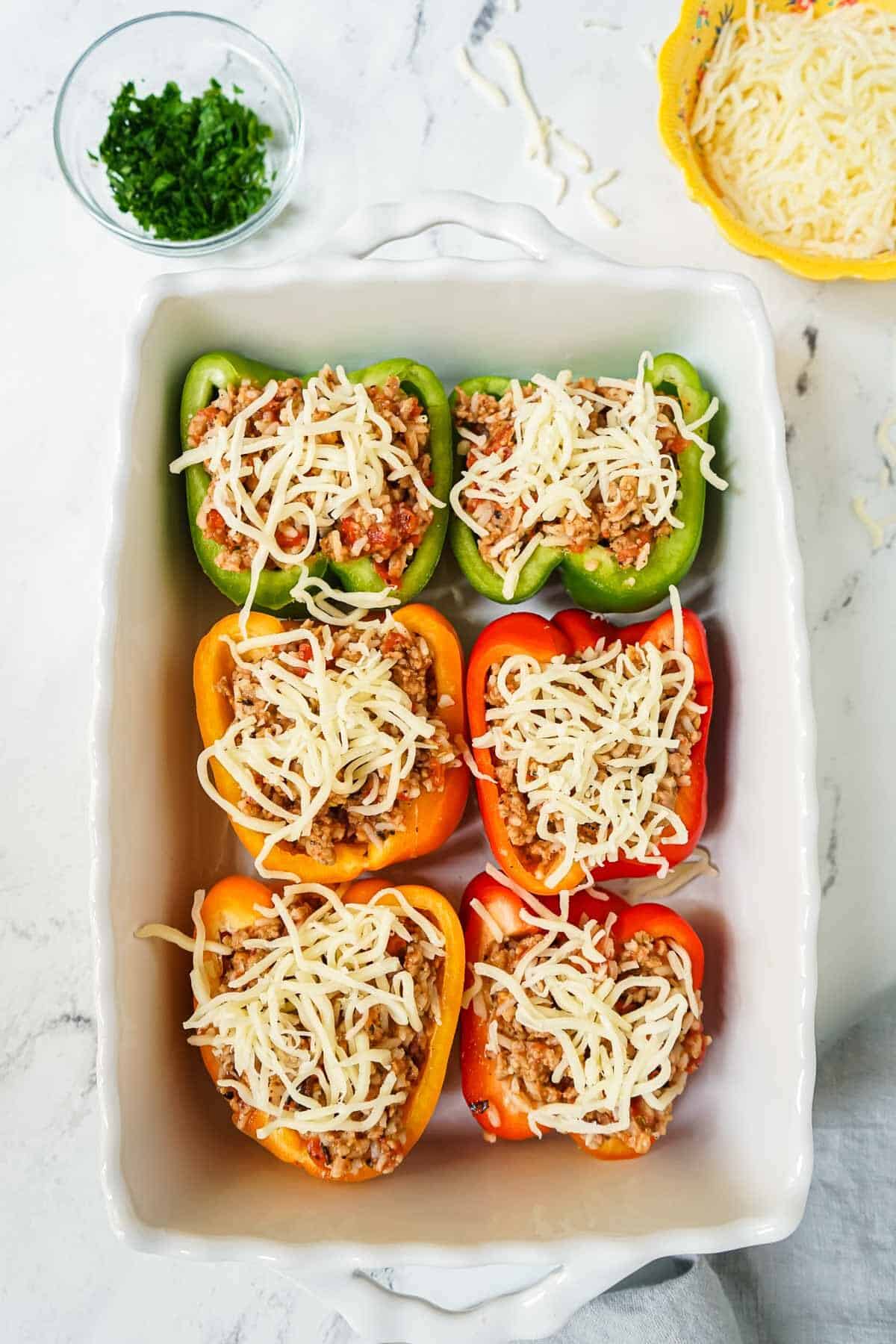 Bell peppers that are filled with stuffing and topped with cheese in a white casserole dish before being cooked.