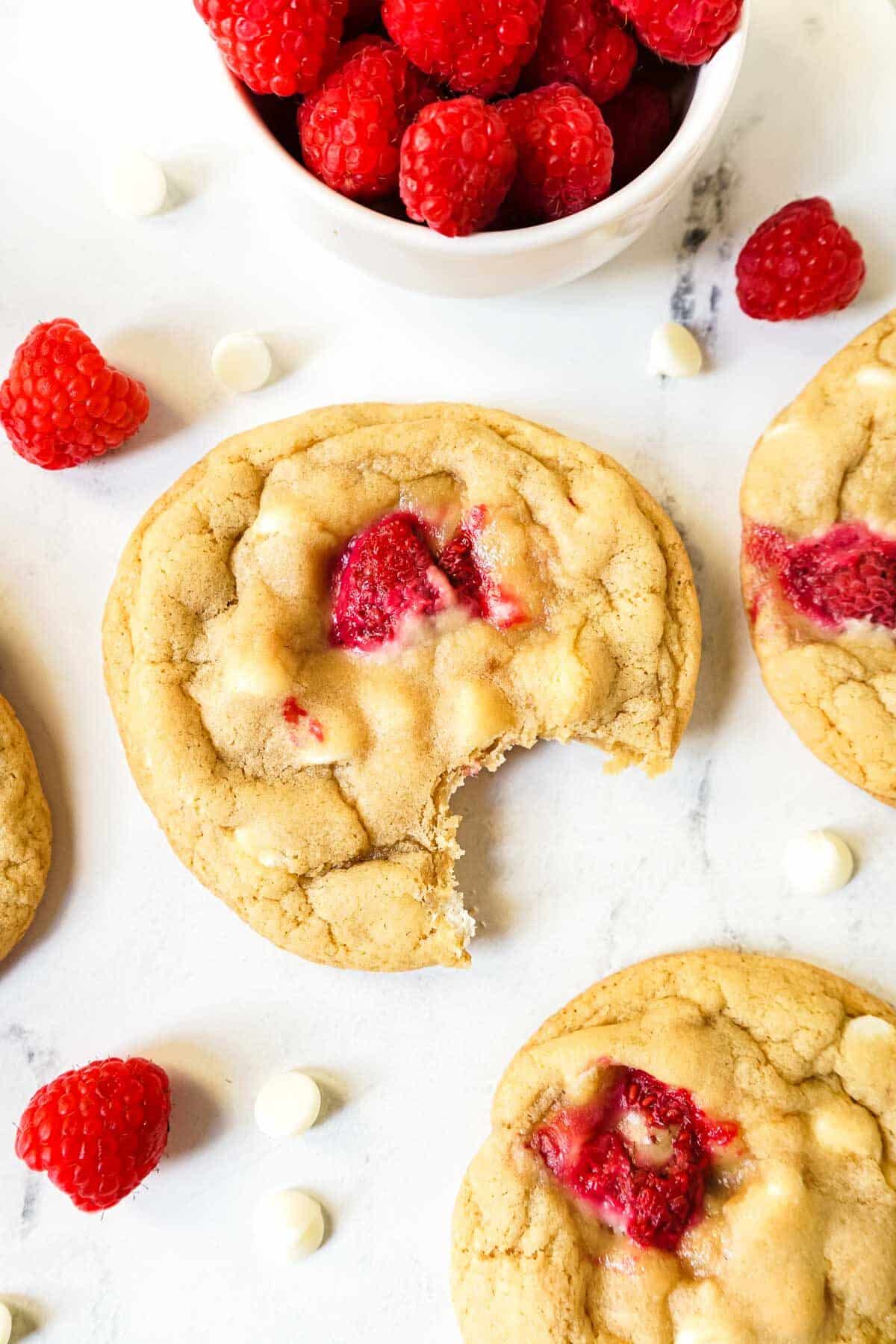 White Chocolate Raspberry cookie with a bite taken out on the counter with white chocolate chips and fresh raspberries.
