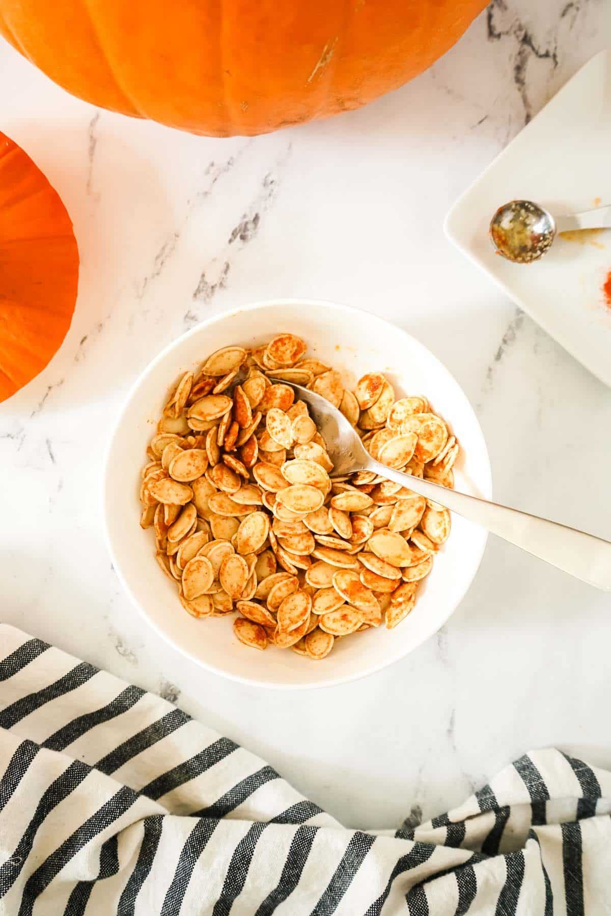 Air fryer pumpkin seeds in a white bowl being tossed with olive oil and spices.