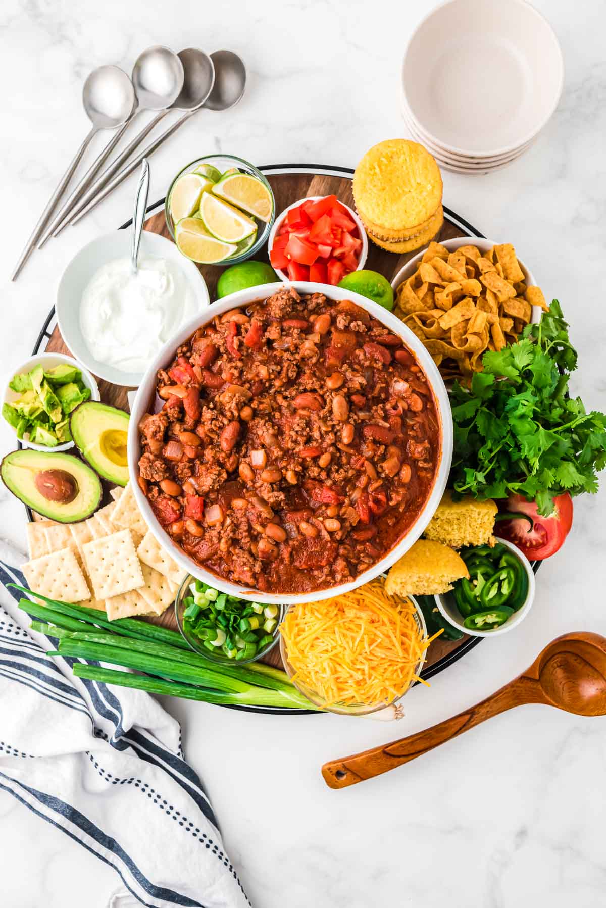 a big white bowl of chili on a wooden chopping block with all of the toppings for chili around it.