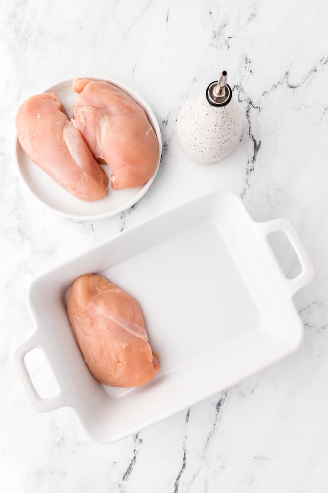 Raw chicken breast in a white baking dish being rubbed with oil