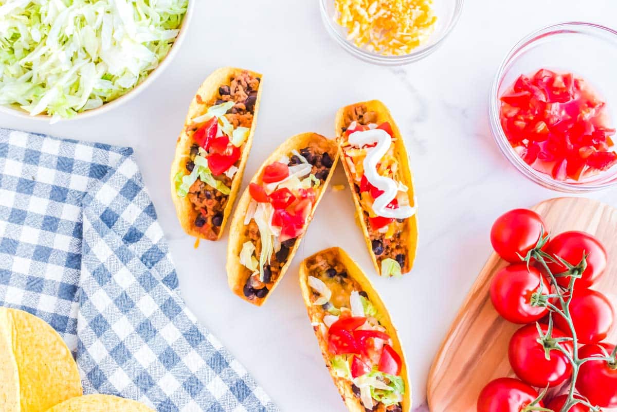Four hard shell ground chicken tacos on a table with tomatoes and taco shells.