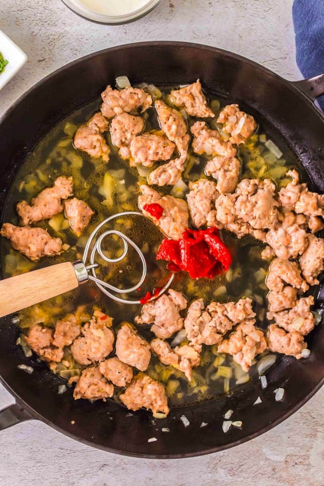 Italian sausage, chicken stock and tomato paste being added to a large skillet.