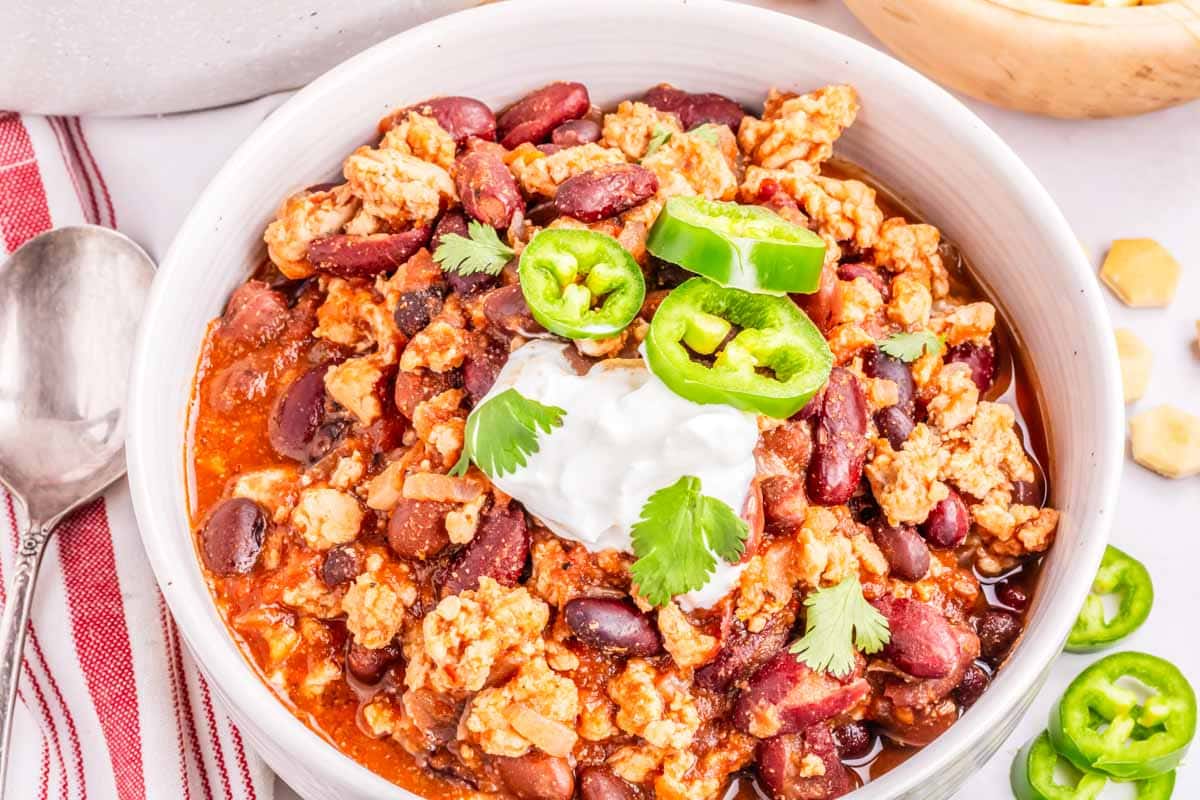 A bowl of ground chicken chili in a white bowl garnished with sour cream and jalapeno slices.