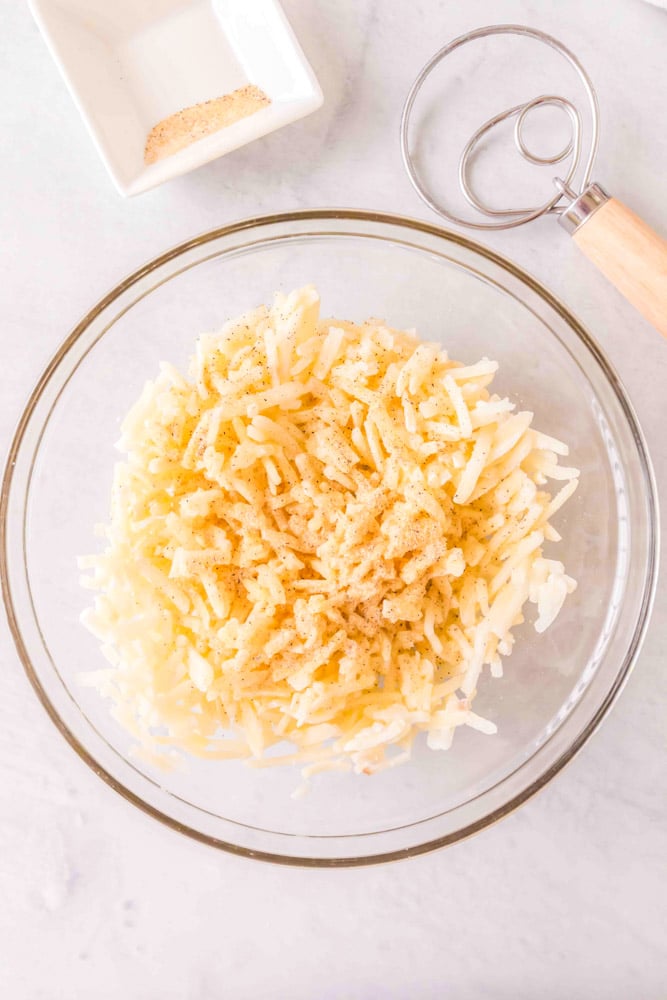 Shredded frozen hash browns in a large glass bowl with seasonings.