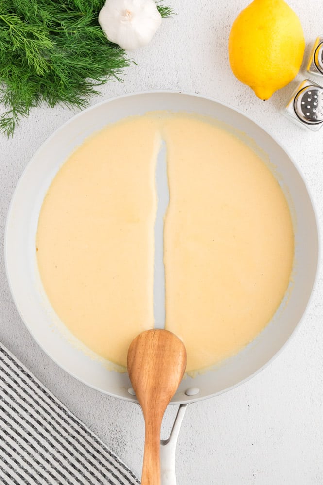 Dill cream sauce in a pan with a wooden spoon to show consistency.