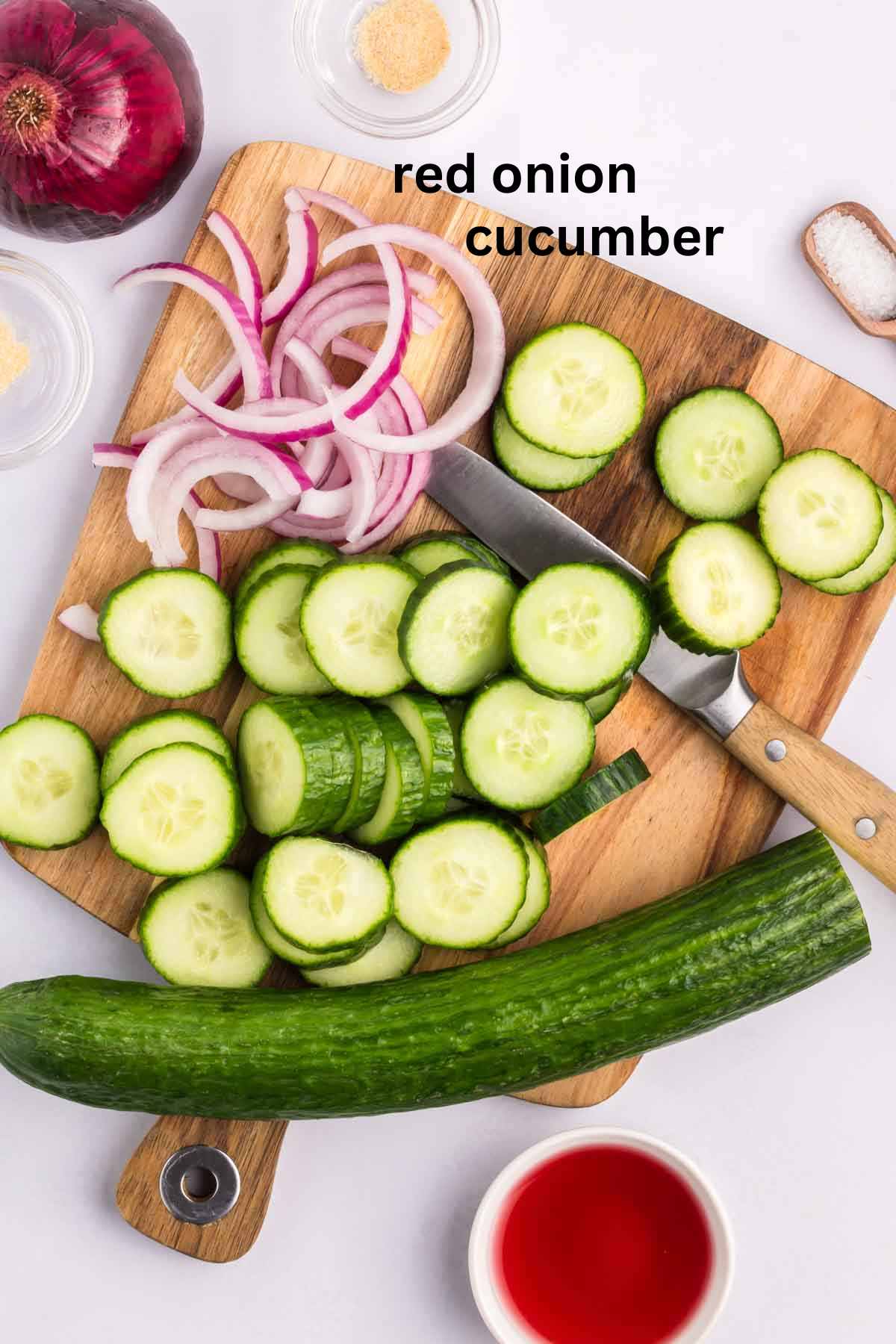 Cucumbers and red onions being chopped on a wooden cutting board.
