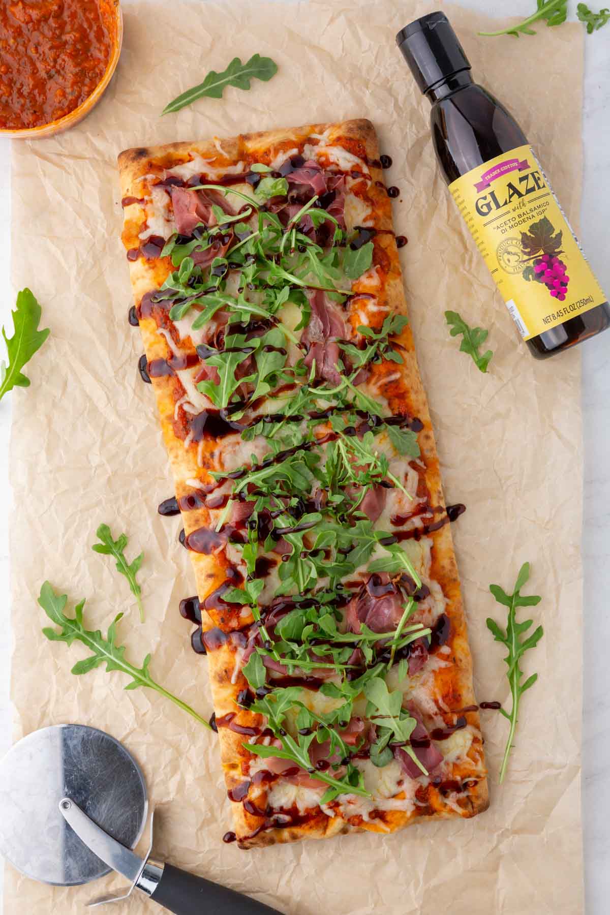 Trader Joe's pizza with balsamic glaze, and arugula on a piece of parchment paper with a pizza cutter.