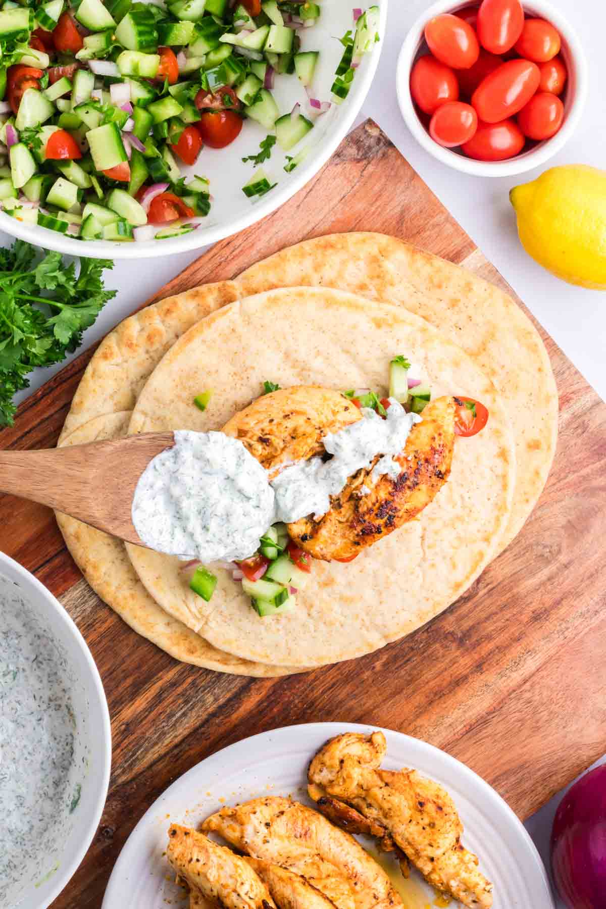 Greek chicken pitas on a cutting board with tzatziki sauce being spooned on them.
