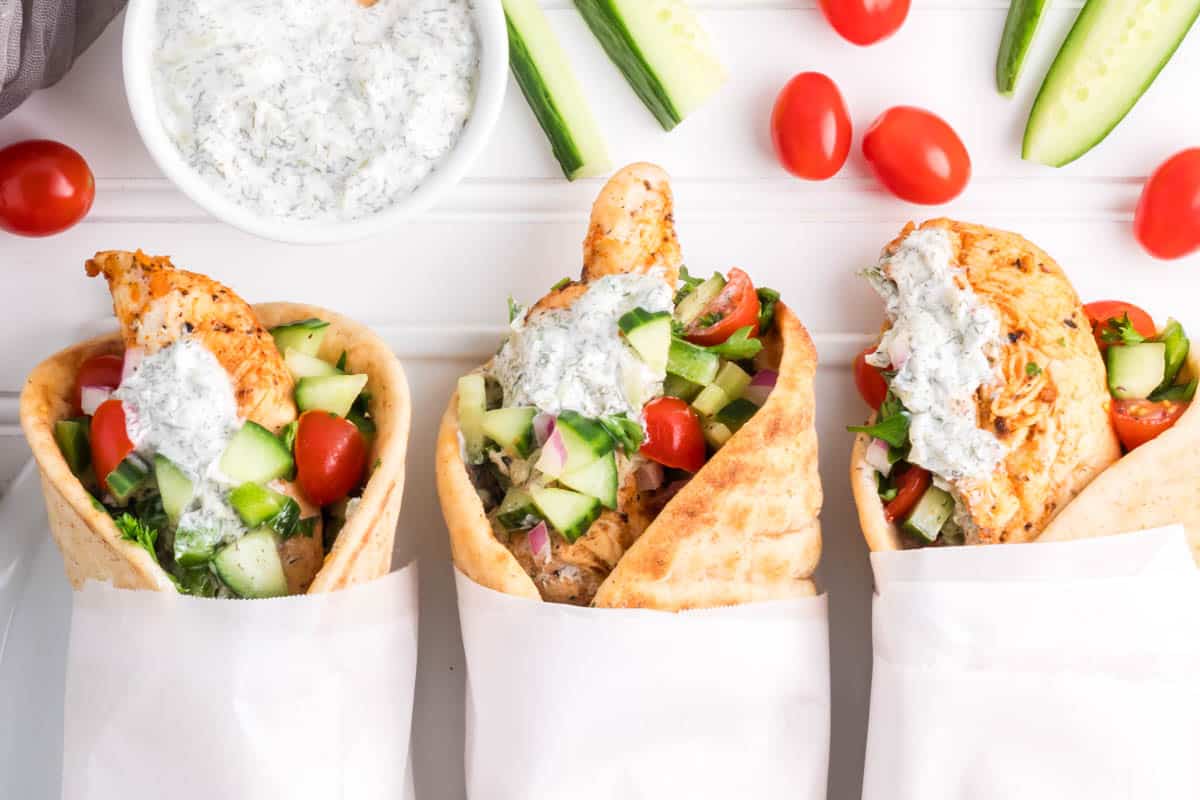 Three Greek chicken pitas wrapped in paper with tzatziki sauce.